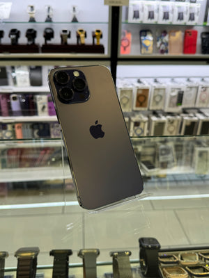 A dark gray Apple iPhone Pro shown from the back, resting on a clear display stand inside a retail store with watch displays in the background.