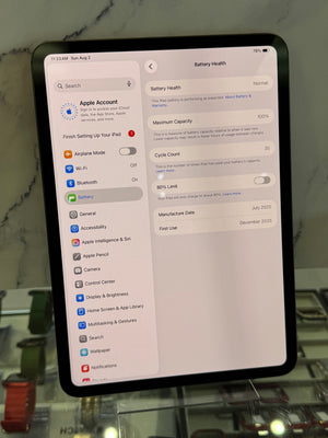 A tablet device displaying the Apple Settings app screen for Battery Health, photographed on a reflective glass surface with blurred background objects.