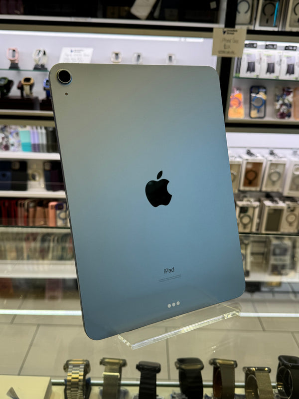 An Apple iPad shown from the back, featuring a light silver-gray finish with the iconic black Apple logo in the center. The device is photographed inside a retail store setting with display shelves and electronic accessories visible in the blurred background.
