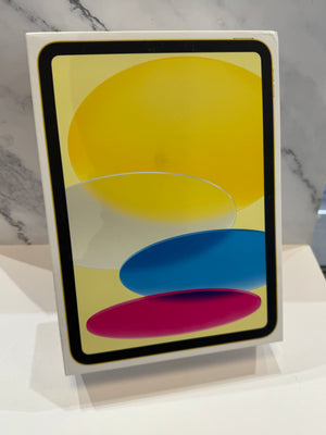 A tablet device in a white retail box, standing upright on a white surface against a light gray marble-patterned background. The screen displays abstract overlapping shapes in yellow, white, blue, and magenta.