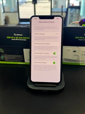 A smartphone displayed upright on a charging stand in a retail or office environment, with a battery health settings screen visible on the phone screen. 