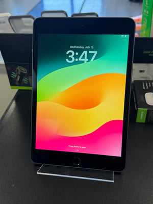 A black tablet, likely an iPad, standing upright on a clear acrylic display stand on a dark tabletop. The screen is on and displays the colorful lock screen wallpaper with the time 3:47.