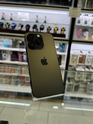 An Apple iPhone in dark bronze/olive green, standing upright on a clear acrylic display stand inside a retail store. The Apple logo is visible on the back of the phone, with blurred store shelves in the background.
