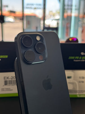 A dark gray Apple iPhone Pro showing the back of the device with its triple-camera system and the Apple logo, photographed in a real-world setting with product packaging boxes blurred in the background.