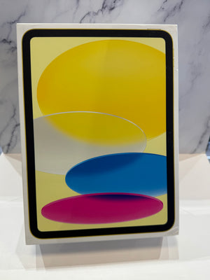 A tablet device in a white retail box, standing upright on a white surface against a light gray marble-patterned background. The screen displays a colorful abstract wallpaper with yellow, blue, pink, and white shapes.