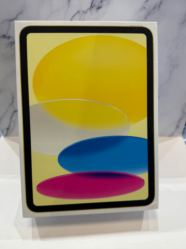 A tablet device in a white retail box, standing upright on a white surface against a light gray marble-patterned background. The screen displays a colorful abstract wallpaper with yellow, blue, pink, and white shapes.