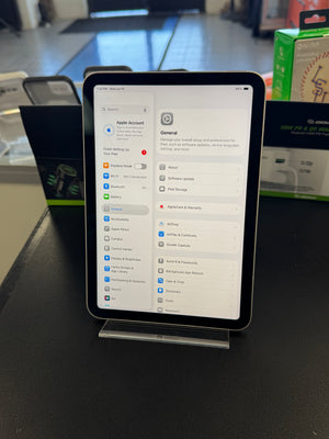 A tablet device with a black bezel and silver side frame, displaying a settings interface on the screen, photographed on a dark tabletop with store items blurred in the background.