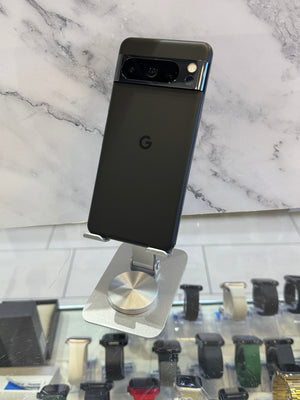 A black Google Pixel smartphone shown from the back, mounted on a silver metal display stand. The phone is positioned in front of a white marble-patterned wall, with a glass display case containing various smartwatches visible in the blurred background.