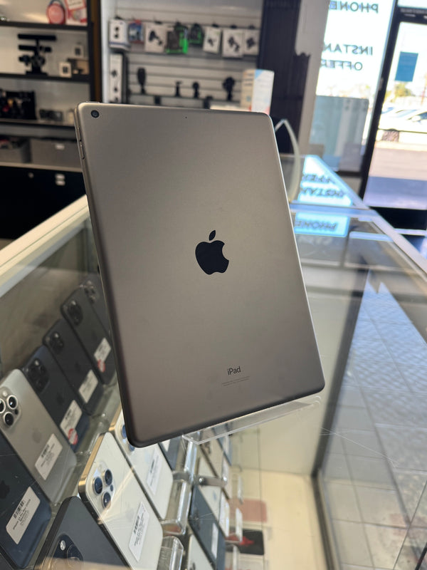 A silver Apple iPad shown from the back, standing on a clear display stand inside a retail store. The Apple logo is visible in the center, with other devices and store fixtures blurred in the background.