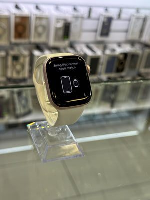 A smartwatch with a black rectangular display and a light beige band, mounted on a clear acrylic stand in a retail display case with blurred Apple products in the background.