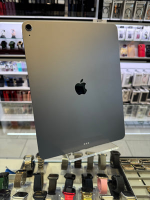A silver Apple iPad shown from the back, featuring the black Apple logo and a small camera lens cutout in the bottom-left corner. The device is photographed inside a retail store setting with shelves of accessories visible in the blurred background.