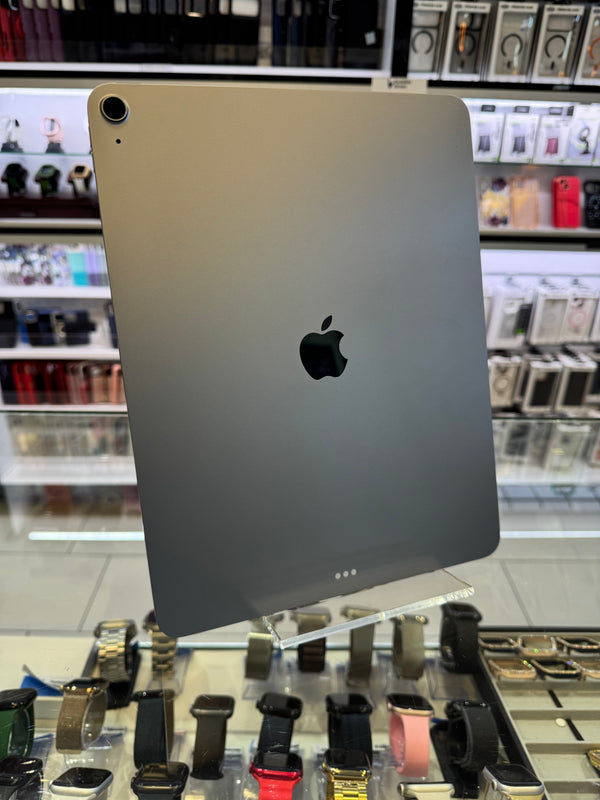 A silver Apple iPad shown from the back, featuring the black Apple logo and a small camera lens cutout in the bottom-left corner. The device is photographed inside a retail store setting with shelves of accessories visible in the blurred background.
