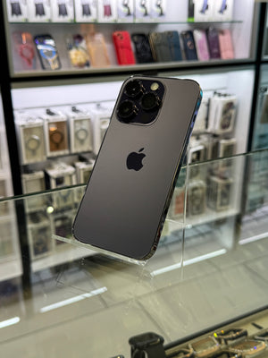 A dark gray Apple iPhone 12 Pro shown from the back, resting on a glass display case inside a retail store. The Apple logo is clearly visible on the back of the device.