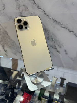 A gold-colored Apple iPhone 12 Pro shown from the back, resting on a silver metal display stand in front of a white and gray marble-patterned wall.