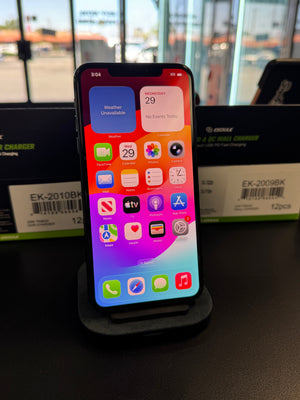 A black smartphone with a colorful home screen wallpaper stands on a black charging dock on a dark tabletop. In the background, two black wall chargers are visible on a store counter.