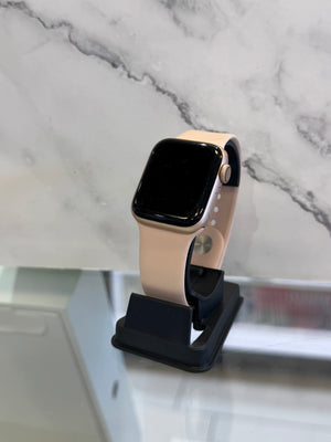 A smartwatch with a light peach-beige sport band and a black screen, displayed on a black charging stand against a white marble-patterned background.