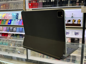 A black tablet case with an attached keyboard and stand, shown standing upright on a glass display counter inside a retail store. The background shows shelves filled with various colorful phone cases.
