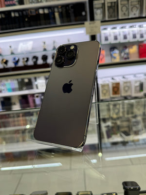 A dark gray Apple iPhone Pro shown from the back, displayed on a clear stand inside a retail store with shelves of electronics in the blurred background.