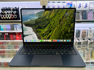 A dark gray Apple MacBook laptop with an open screen displaying a lush green landscape wallpaper, set on a glass counter inside a retail store with shelves of accessories in the background.