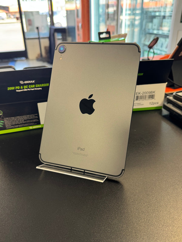 A silver Apple iPad shown from the back, standing on a small display stand on a dark tabletop. The Apple logo is visible in the center, with retail packaging and a store interior blurred in the background.