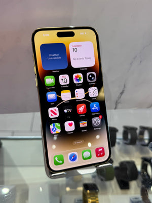 A black iPhone with a gold-toned edge, displaying its home screen on a glass display stand in a store setting.