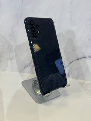 Samsung smartphone shown from the back in a dark navy blue color, resting on a silver metal display stand against a white marble-patterned background.