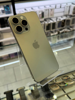 An Apple iPhone with a light gold finish, shown from the back and resting on a clear acrylic display stand inside a retail store.