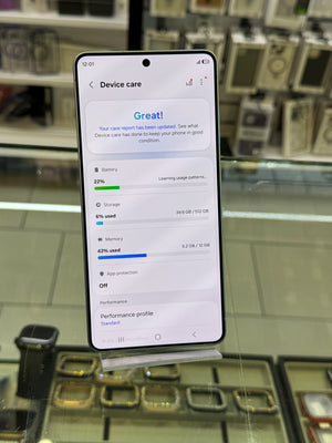 A smartphone displayed upright on a stand in a retail setting, showing a device care screen with battery, storage, and memory status. The phone has a black front and is photographed through a glass display case with blurred electronics in the background.