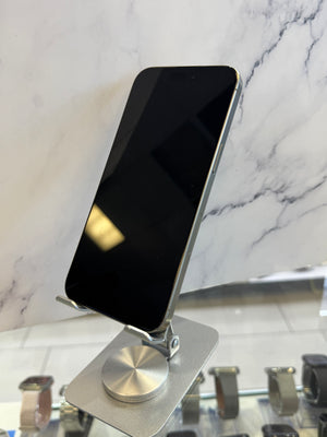 A black smartphone with a silver metallic edge stands on a small silver display stand. The phone is photographed indoors against a white marble-patterned wall, with watch displays visible along the bottom edge of the frame.