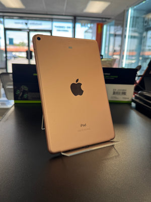 A gold Apple iPad standing upright on a clear display stand on a dark countertop, with a blurred store interior in the background.