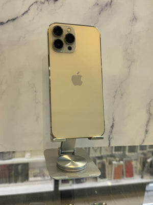 A gold-colored Apple iPhone, likely an iPhone 12 Pro or 13 Pro model, shown from the back resting on a white marble-patterned surface with a metal stand visible on the right.