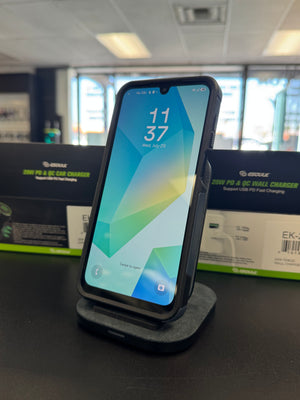 A black smartphone in a protective case stands upright on a charging dock on a dark tabletop. The phone screen is on, showing a colorful geometric wallpaper with the time 11:37.