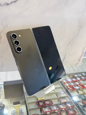 A black Samsung Galaxy S24 Ultra smartphone shown from the back, standing on a display stand in a retail or showroom setting with a marble-patterned backdrop and watch displays below.