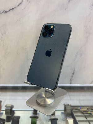 A dark blue-gray Apple iPhone 12 Pro displayed upright on a silver stand, showing the back of the phone with the Apple logo and triple camera module against a marble-patterned background.