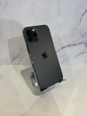 A dark gray Apple iPhone 12 Pro shown from the back, resting on a silver metal stand against a white marble-patterned background.