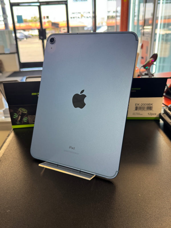 A silver Apple iPad Pro shown from the back, standing on a clear display stand on a dark tabletop. Apple branding and the iPad name are visible on the device, with retail packaging boxes in the background inside a store.