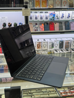 A dark gray laptop with a black screen and keyboard is displayed on a glass counter inside a retail store, with shelves of phone cases and accessories visible in the background.
