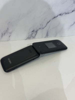 A black flip phone with a singular camera and a small screen.  It is resting on a white tabletop.