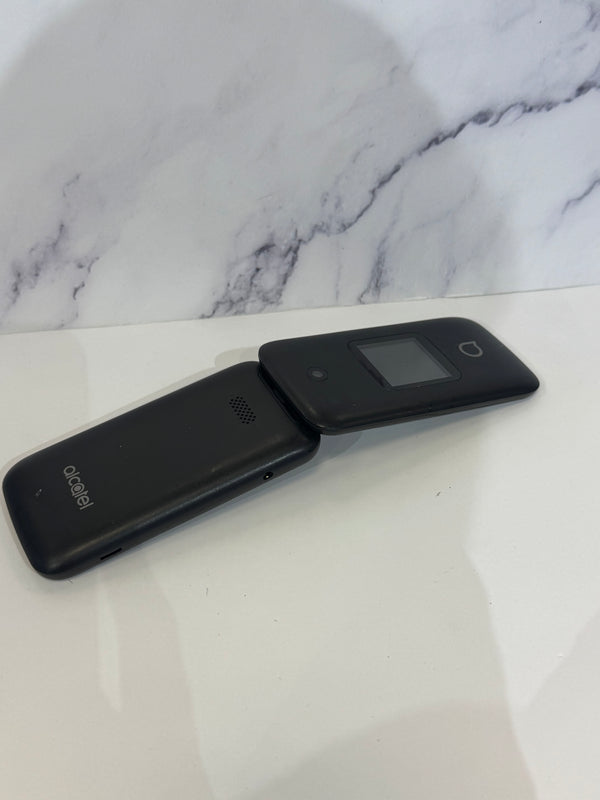 A black flip phone with a singular camera and a small screen.  It is resting on a white tabletop.