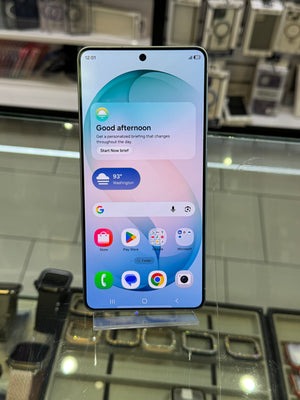 A smartphone with a large front-facing display showing a home screen, standing upright on a clear display stand inside a retail store. Watch straps and shelving are visible in the background.