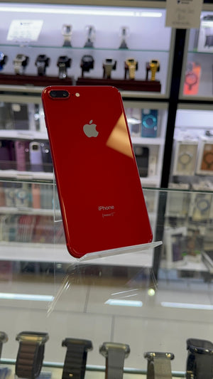 A red Apple iPhone 7 Plus shown from the back, resting on a clear display stand inside a retail store. The Apple logo and 'iPhone' branding are visible on the glossy red backplate.