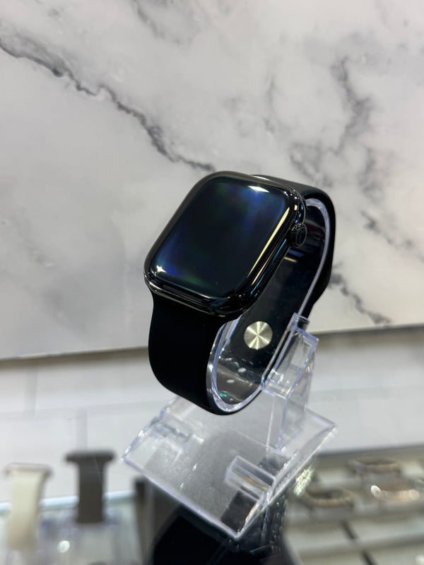 A black smartwatch with a square screen and black band, displayed on a clear acrylic stand against a white marble-patterned background.