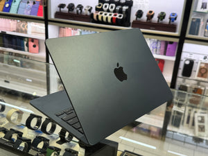 A closed Apple MacBook laptop in dark gray sits on a glass display counter inside a retail store, with shelves of watches and accessories blurred in the background.