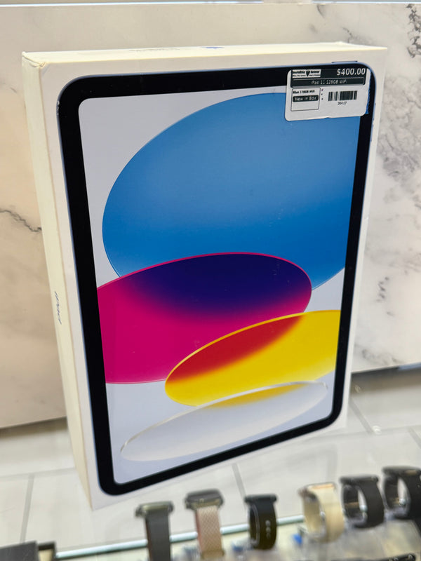 A boxed iPad shown in its retail box, with a colorful abstract screen graphic and a price sticker in the top right corner. The box is photographed indoors against a light marble-patterned surface, with watch straps visible along the bottom edge.