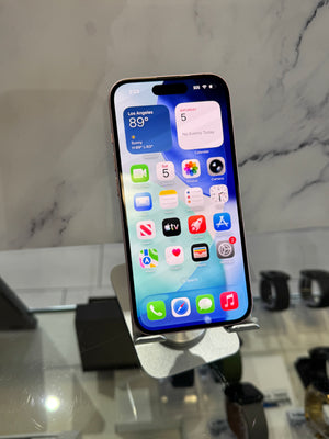 A black iPhone with a silver metallic frame, displaying its home screen on a small metal stand. The phone is set against a blurred background of a marble-patterned wall and a glass display counter.