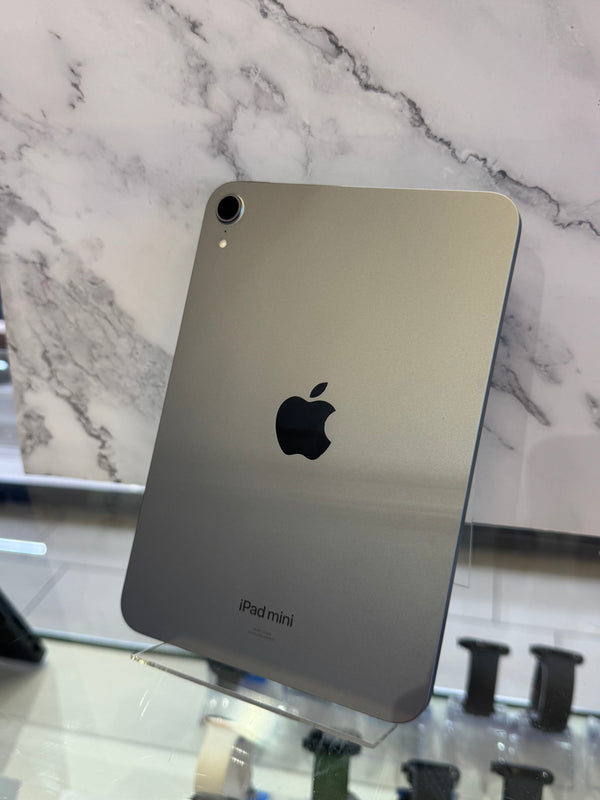 An Apple iPad mini in metallic silver, showing the back of the device with the Apple logo and 'iPad mini' text, displayed on a clear stand in front of a marble-patterned background.