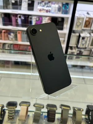 A black Apple iPhone 11 standing upright on a clear acrylic display stand inside a retail store. The back of the phone is visible, showing the Apple logo, with blurred shelves of other phones and smartwatches in the background.