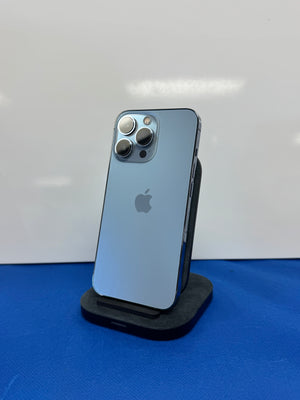 An Apple iPhone Pro in blue-gray color, standing upright on a black charging dock against a white background with a blue surface below.
