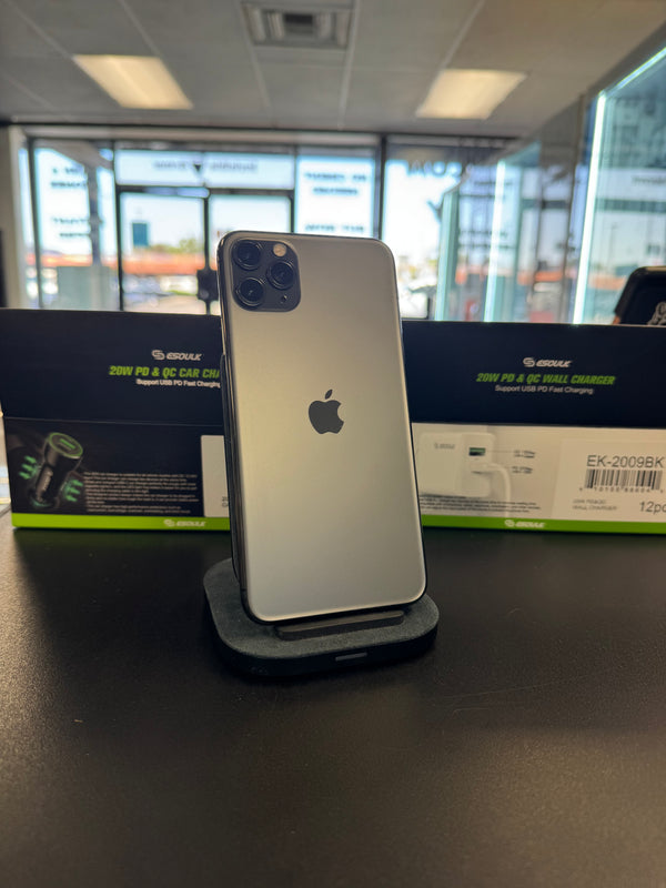 A silver Apple iPhone stands upright on a black charging dock on a dark tabletop, with a black and green Apple 25W PD & 60W Wall Charger box behind it in a retail or office setting.