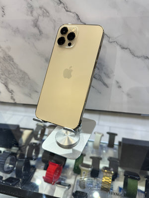 A gold Apple iPhone 12 Pro displayed on a silver stand, showing its back panel with the Apple logo and triple rear cameras, set against a marble-patterned background.